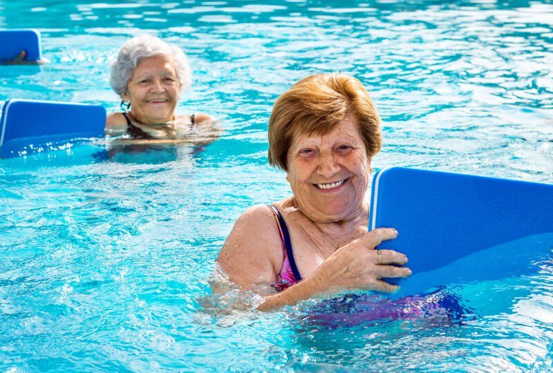 Two aged ladies holding surf board in the swimming pool